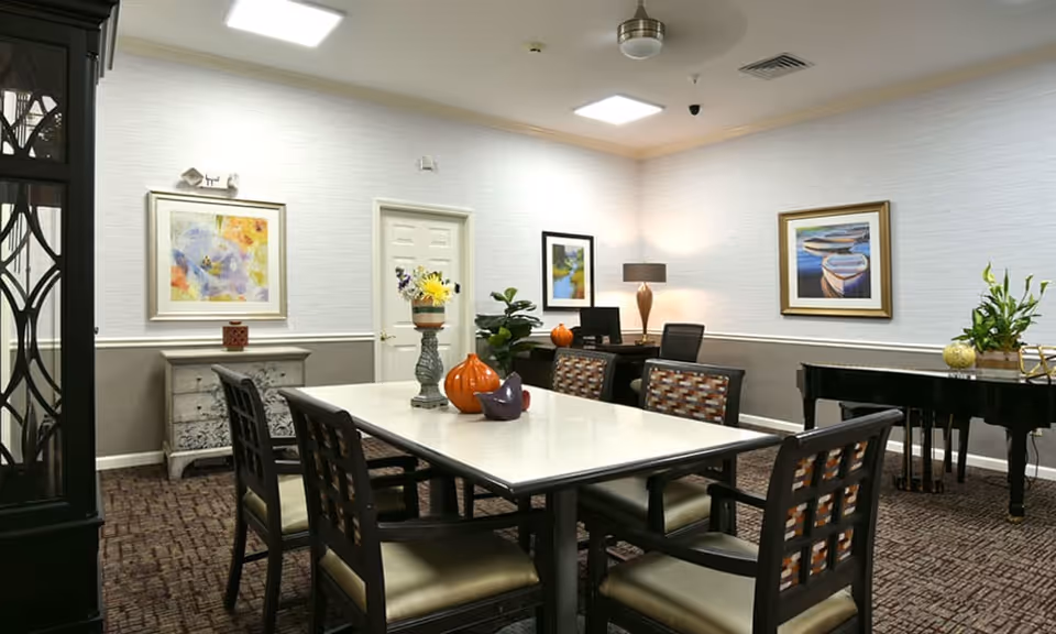 A well-lit interior room with a rectangular table surrounded by six chairs. The table has decorative items including a vase with flowers and ornamental pumpkins. The room features framed artwork on the walls, a black grand piano with a plant on top, a desk with a computer and lamp, and a patterned carpeted floor.