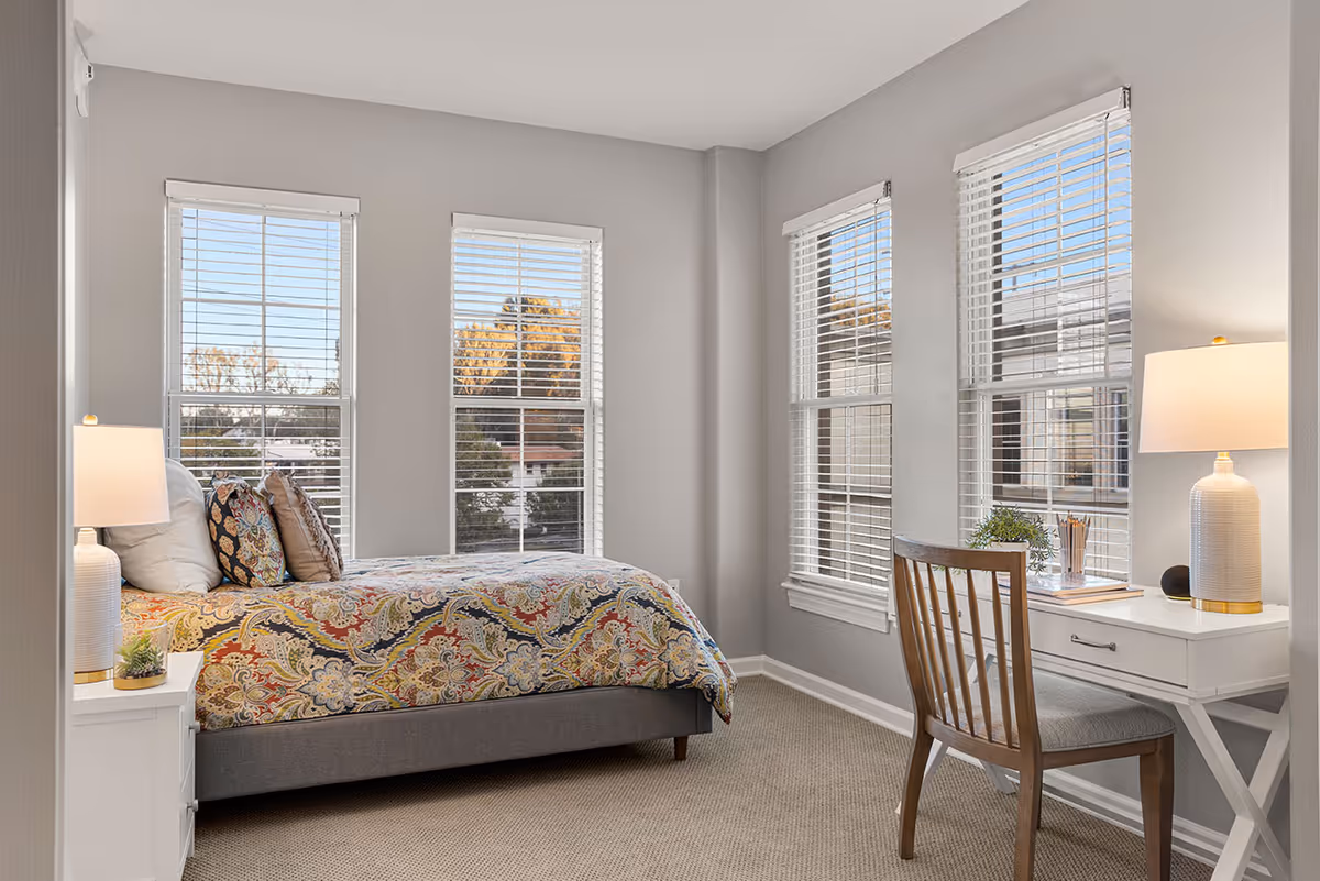 A bright bedroom with three large windows featuring white blinds. The room has a bed with a colorful patterned comforter and several pillows. There is a white nightstand with a lamp and a small plant next to the bed. Opposite the bed is a white desk with a wooden chair, a lamp, a small plant, and some pencils in a holder. The walls are painted light gray and the floor is carpeted.
