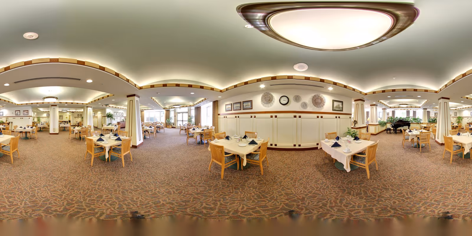 Large assisted-living dining room with neatly set tables and chairs, overhead lighting, and a piano at the far end.