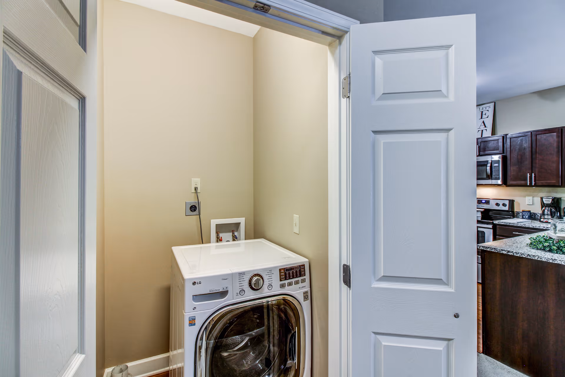 A small laundry area with a white front-loading LG washing machine inside a closet with beige walls and white trim. The closet door is open, revealing part of a kitchen with dark wood cabinets, a microwave, and a granite countertop in the background.