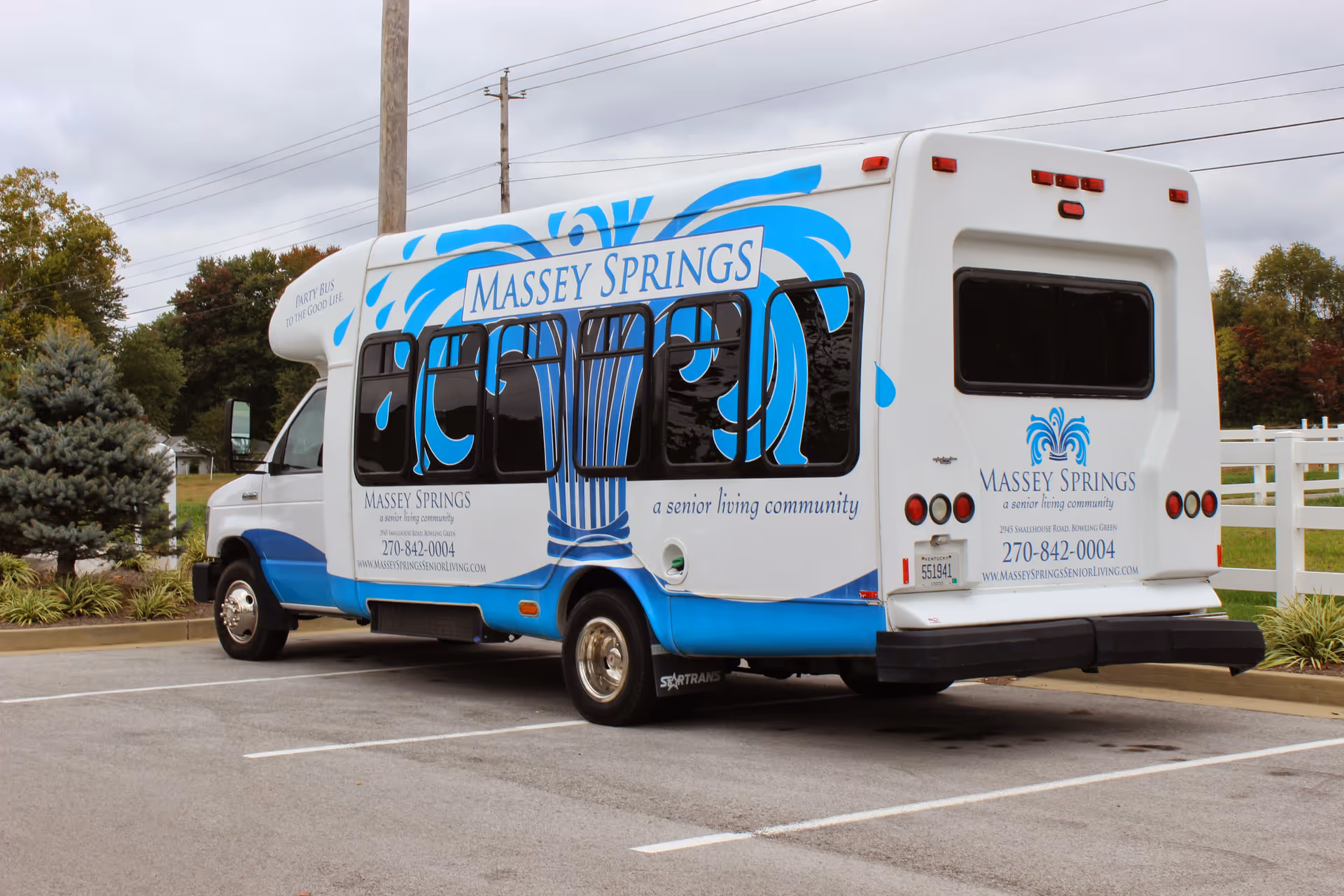 A white and blue shuttle bus parked in a parking lot with the branding 'Massey Springs, a senior living community'. The bus features a large blue water fountain graphic and contact information including a phone number and website. Trees and a white fence are visible in the background under a cloudy sky.