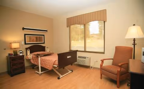 A hospital-style patient bedroom with a bed, nightstand, chair, and window.