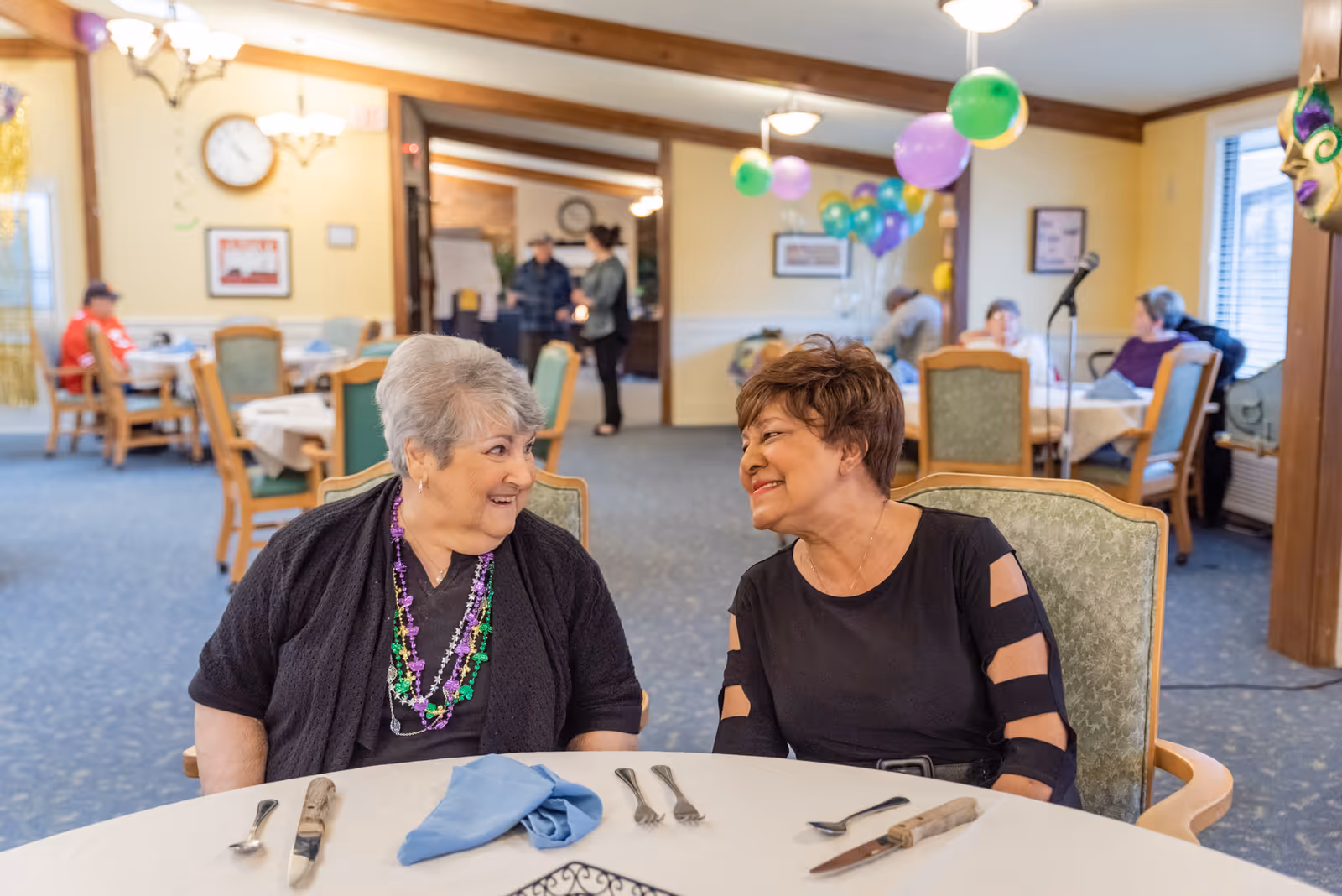 Two elderly women sitting at a round dining table in a decorated room, smiling and engaging in conversation. The table is set with utensils and a folded blue napkin. In the background, other elderly people are seated at tables, and colorful balloons are hanging from the ceiling. The room has warm lighting and a cozy atmosphere.
