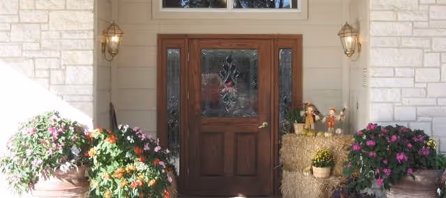Front entrance of a building with a wooden door featuring decorative glass panels, flanked by two wall-mounted lantern-style lights. There are large flower pots with blooming plants on either side of the door, and a small display with hay bales and decorative scarecrows on the right side.