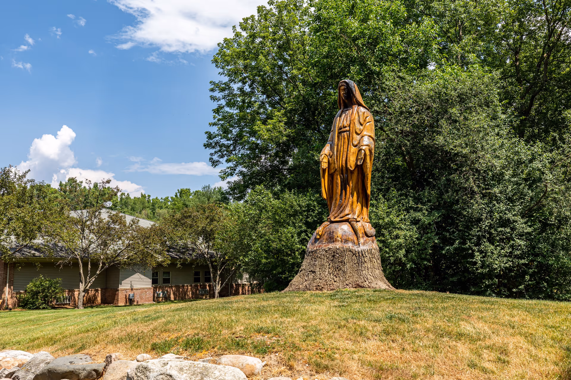 A wooden statue of a robed figure standing on a tree stump in a grassy area with trees and a building in the background under a partly cloudy sky.