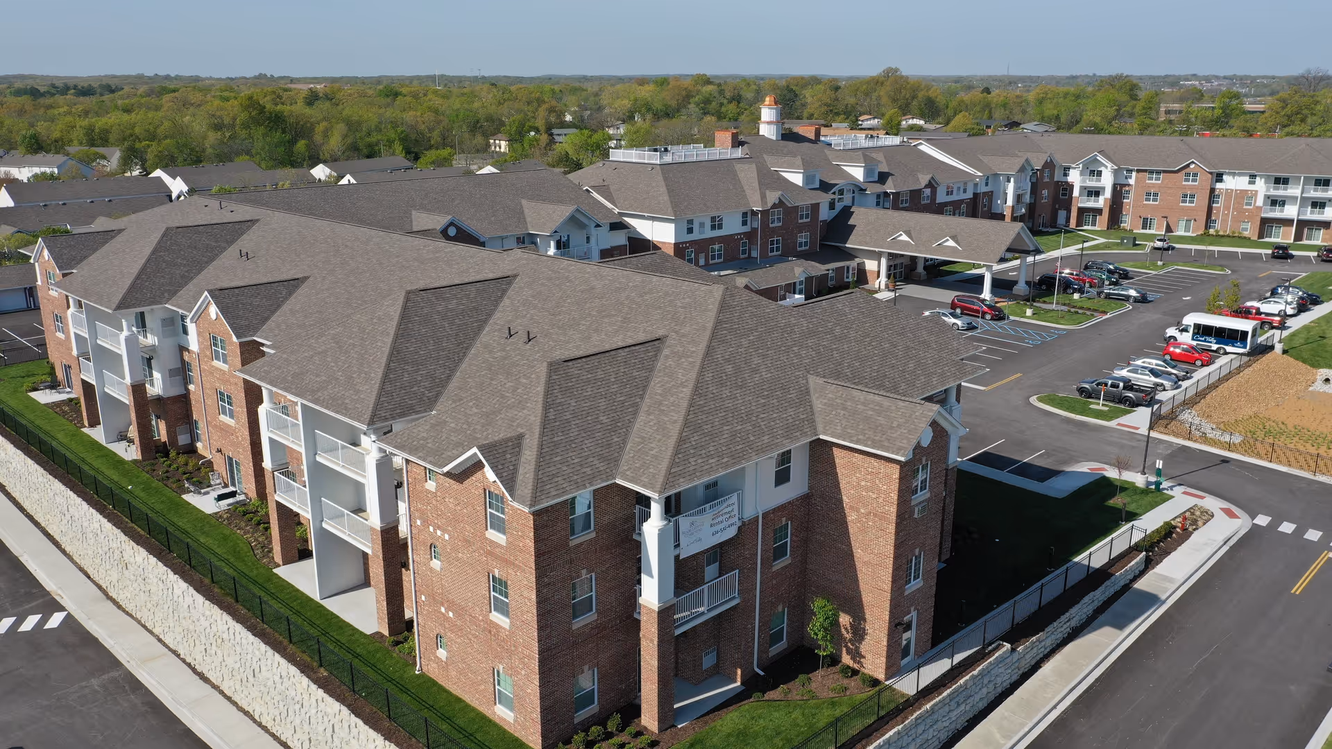 Aerial view of a multi-story brick retirement community complex with parking lot, walkways, and landscaped grounds.