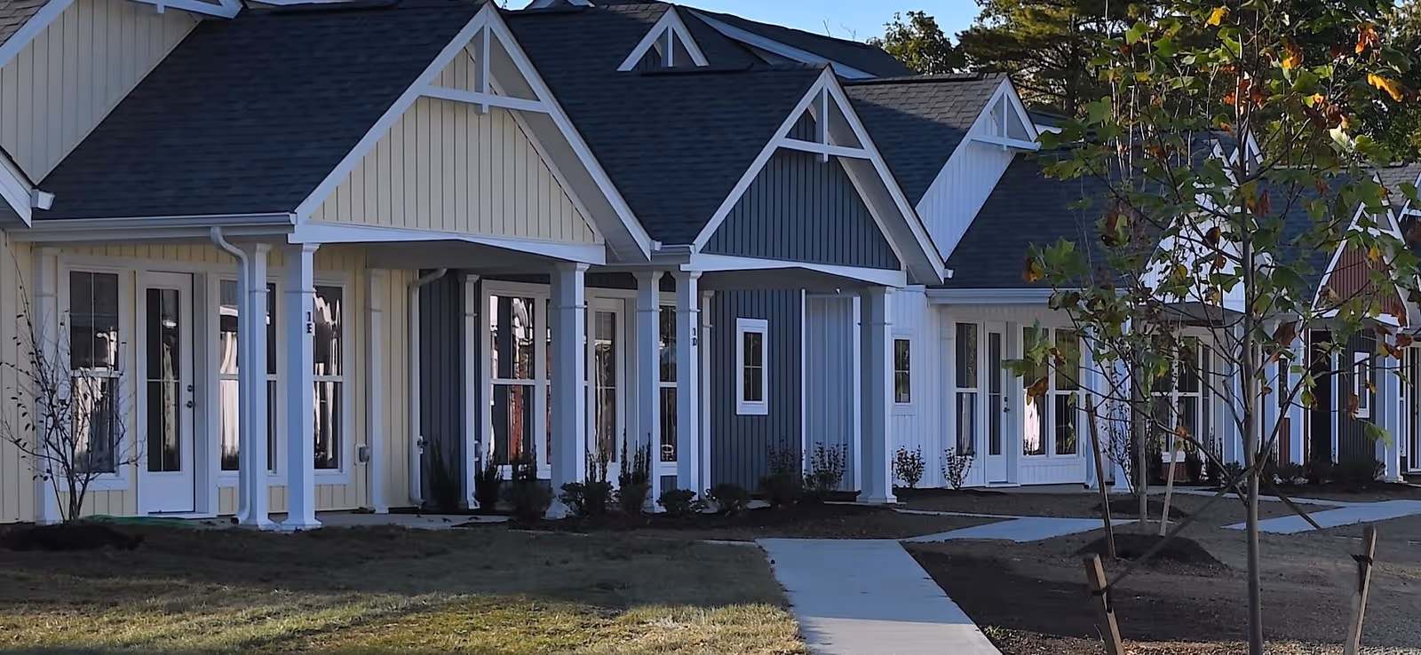 Row of single-story cottage-style units with covered porches, white columns, and a sidewalk in front.
