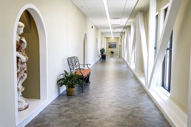 A bright hallway with large windows on the right side supported by white structural beams. On the left side, there are arched wall niches, one containing a decorative statue. A metal bench with a cushion and a potted plant are placed along the left wall. The floor has a patterned tile design, and the hallway extends into the distance with additional plants and a framed picture on the far wall.