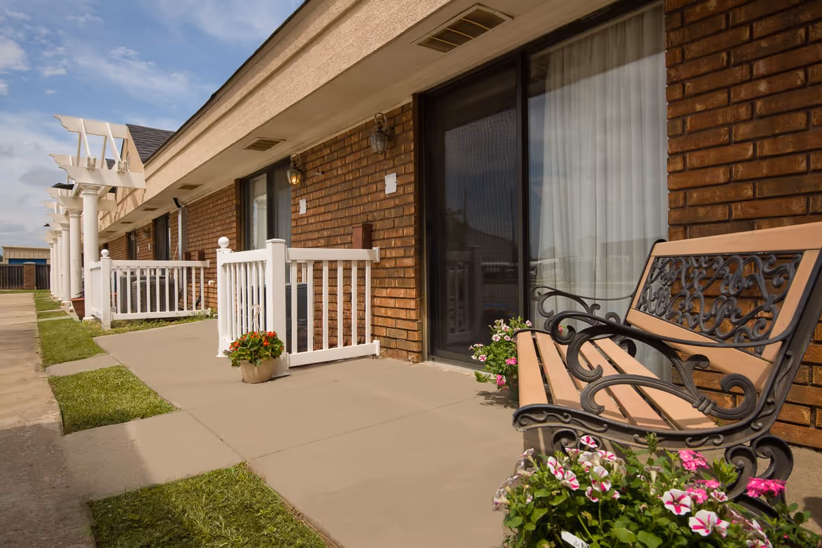 Outdoor patio area of a brick building with white railings, a decorative bench, potted flowers, and sliding glass doors under a partly cloudy sky.