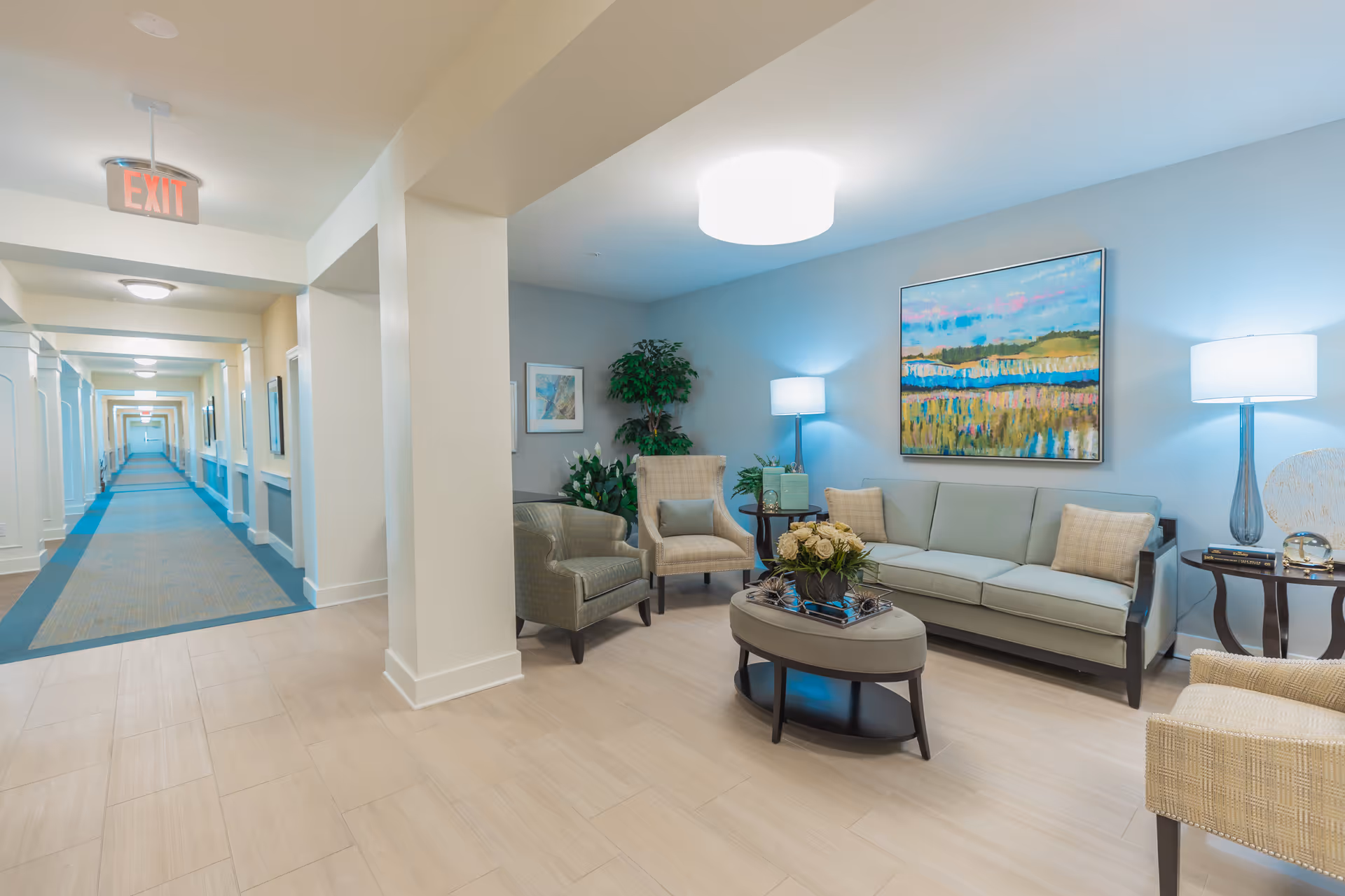 A bright and spacious senior living facility common area with a light blue wall, a comfortable sofa, two armchairs, a round coffee table with a flower arrangement, two side tables with lamps, and a colorful landscape painting. To the left, a long hallway with carpeted flooring and multiple doors extends into the distance, illuminated by ceiling lights and an exit sign.