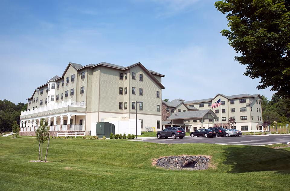 Exterior view of a senior living facility named Braemar at Wallkill, showing two large multi-story buildings with beige siding and gray roofs. There is a parking lot with several cars and an American flag flying near the entrance. The foreground features a well-maintained grassy area with a small tree and a drainage area with rocks.