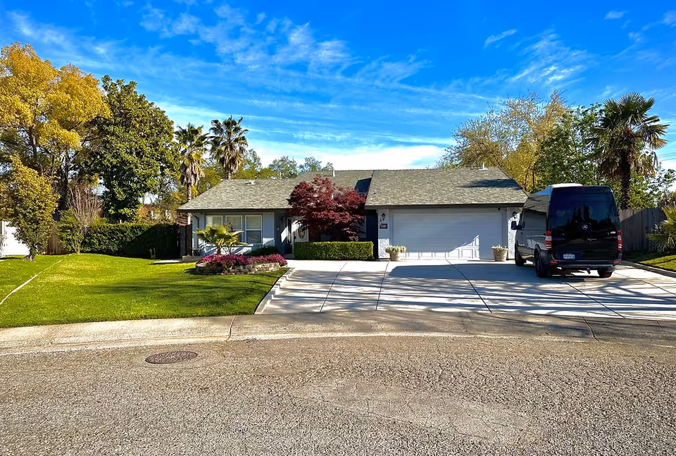 A single-story residential-style senior care facility front with a driveway, trimmed lawn, shrubs, and a parked van.
