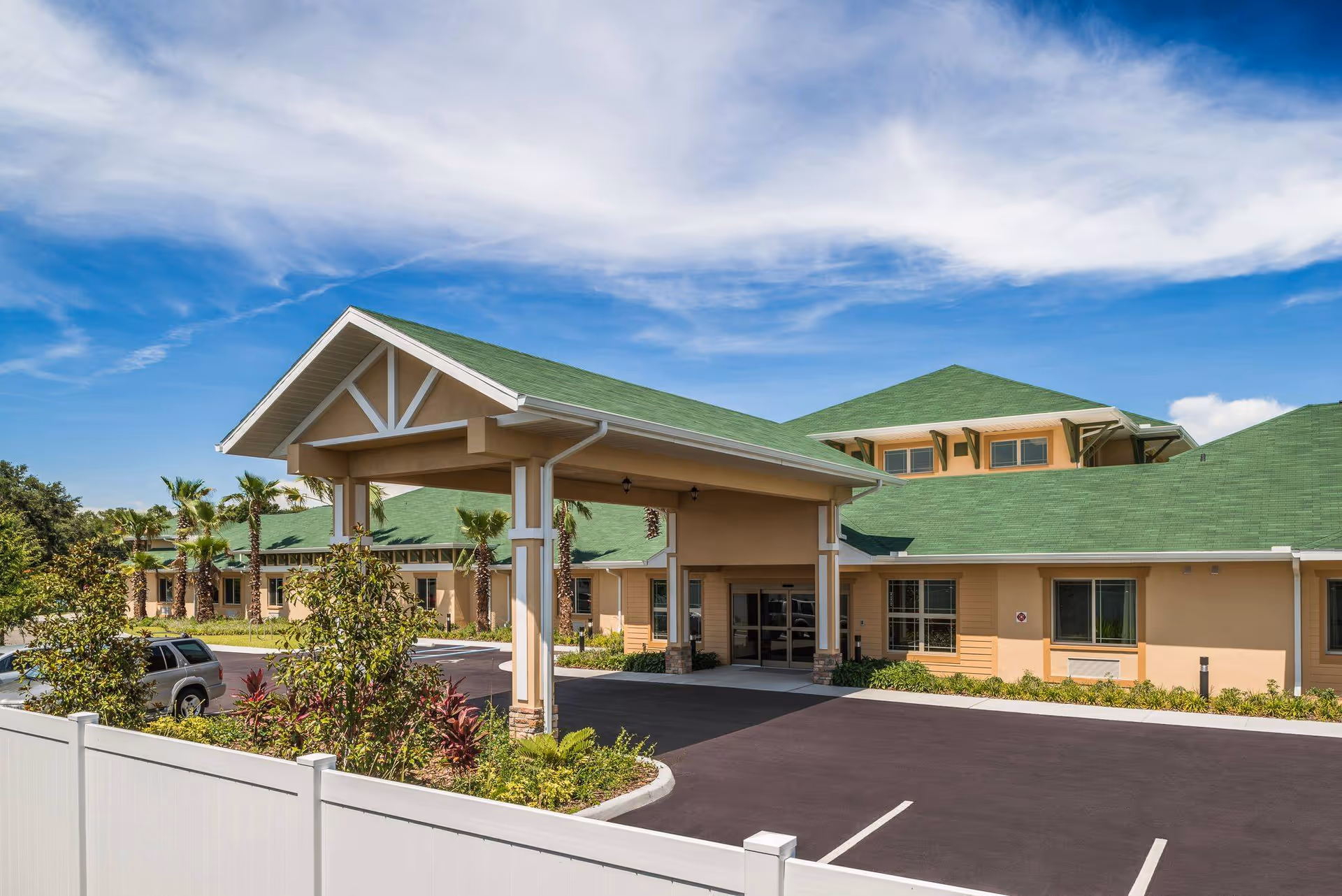Exterior view of a single-story building with a green roof and beige walls under a partly cloudy blue sky. The building has a covered entrance with pillars and a parking area in front. There are palm trees and shrubs around the building.