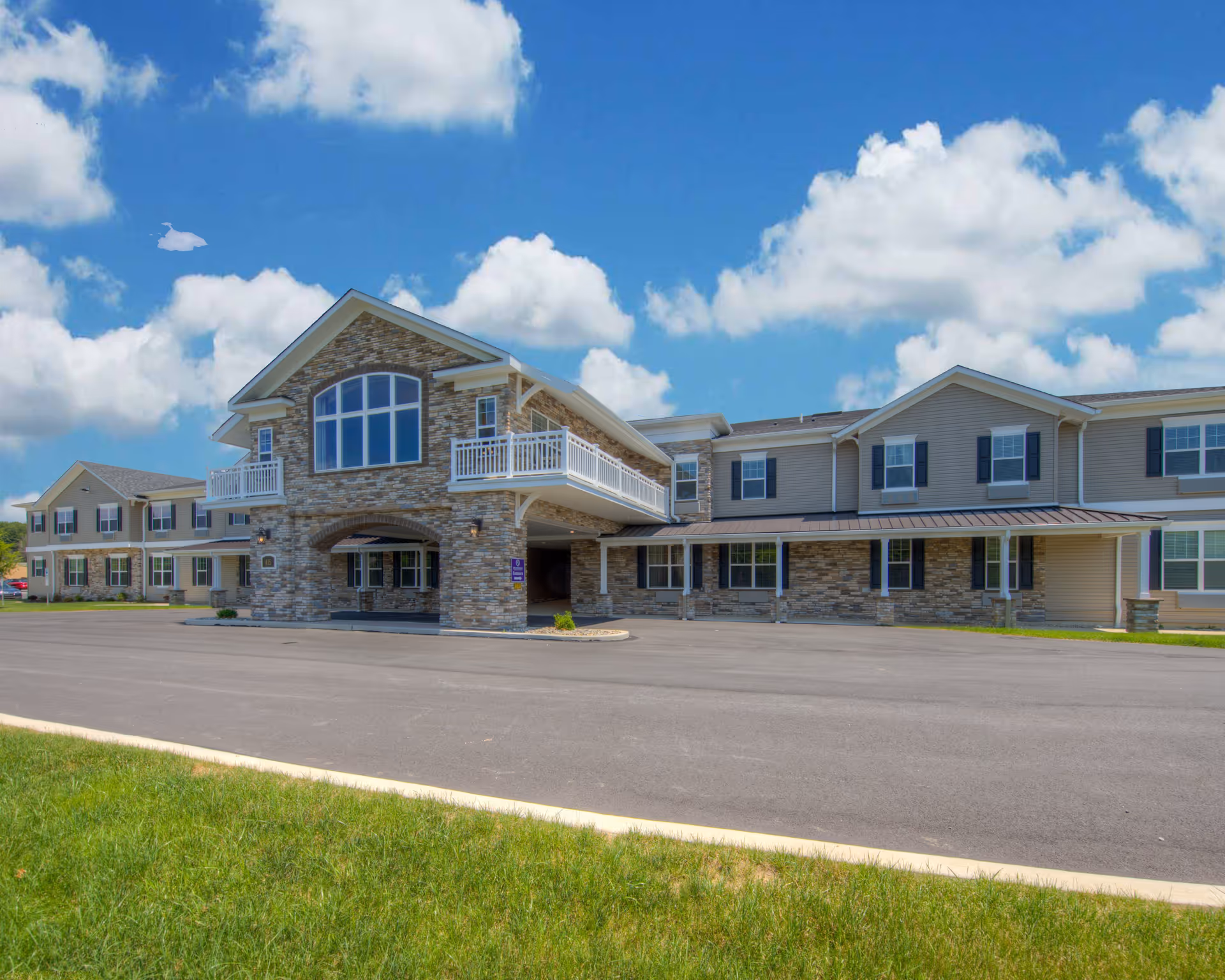 Exterior view of a two-story senior living facility building with stone and siding facade, large windows, balconies, and a covered entrance under a blue sky with scattered clouds. There is a paved driveway and green grass in the foreground.