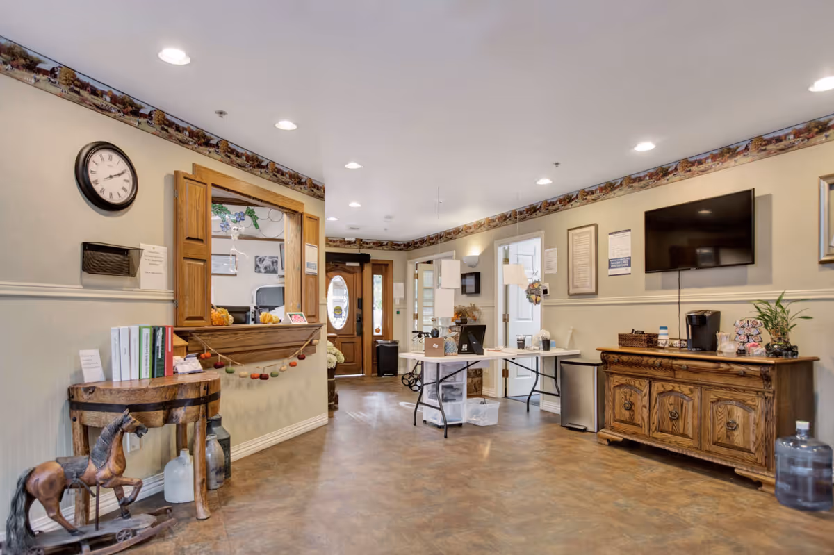 Interior view of a senior living facility reception or common area with a wooden front desk window, a round wooden table with books and a decorative horse figurine, a wall clock, a flat-screen TV mounted on the wall, and a wooden cabinet with a coffee maker and plants. The room has beige walls with a decorative border near the ceiling and recessed lighting.