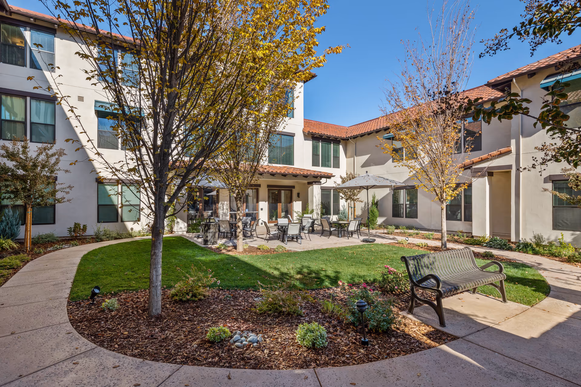 Outdoor courtyard area of a senior living facility with a curved concrete walkway, green grass, small trees, and landscaped flower beds. There are several patio tables with umbrellas and chairs, as well as a metal bench. The building surrounding the courtyard has multiple windows and a tiled roof under a clear blue sky.