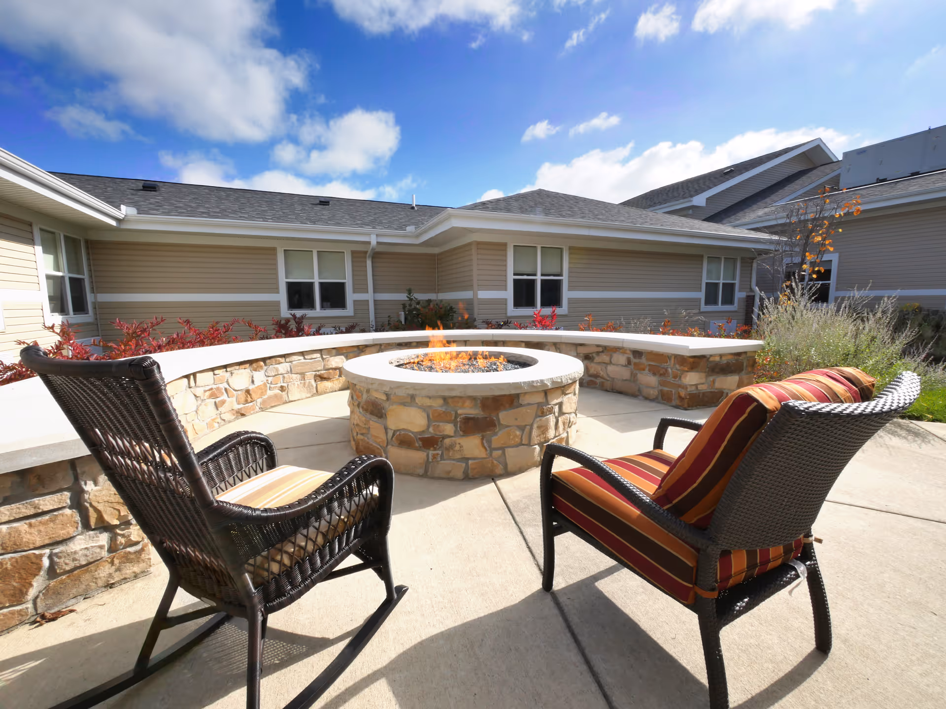 Outdoor patio area at Aperion Care Marion featuring a circular stone fire pit with flames, surrounded by a curved stone bench and two cushioned wicker chairs under a partly cloudy blue sky.