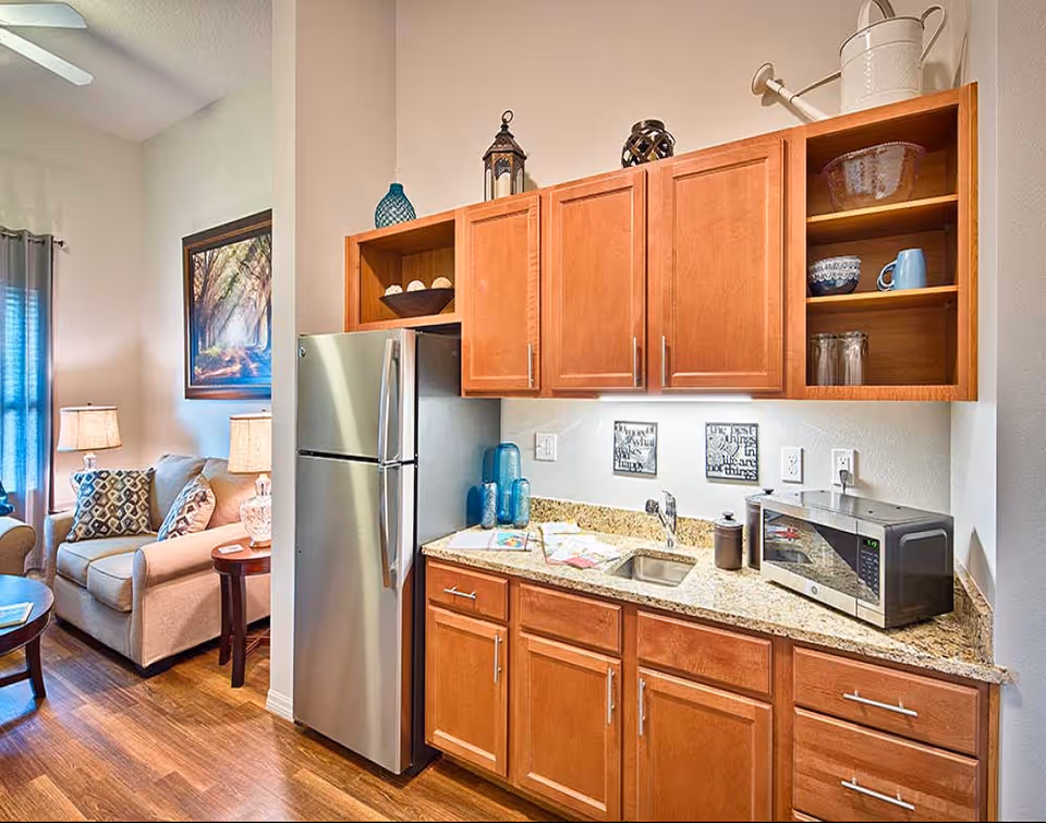 A bright and cozy kitchen area with wooden cabinets, a granite countertop, a stainless steel refrigerator, a microwave, and a small sink. Decorative items such as blue glass bottles, a bowl with decorative balls, and a watering can are placed on the cabinets and countertop. In the background, a living room area is visible with a beige sofa, patterned pillows, a side table with a lamp, and a framed nature artwork on the wall.