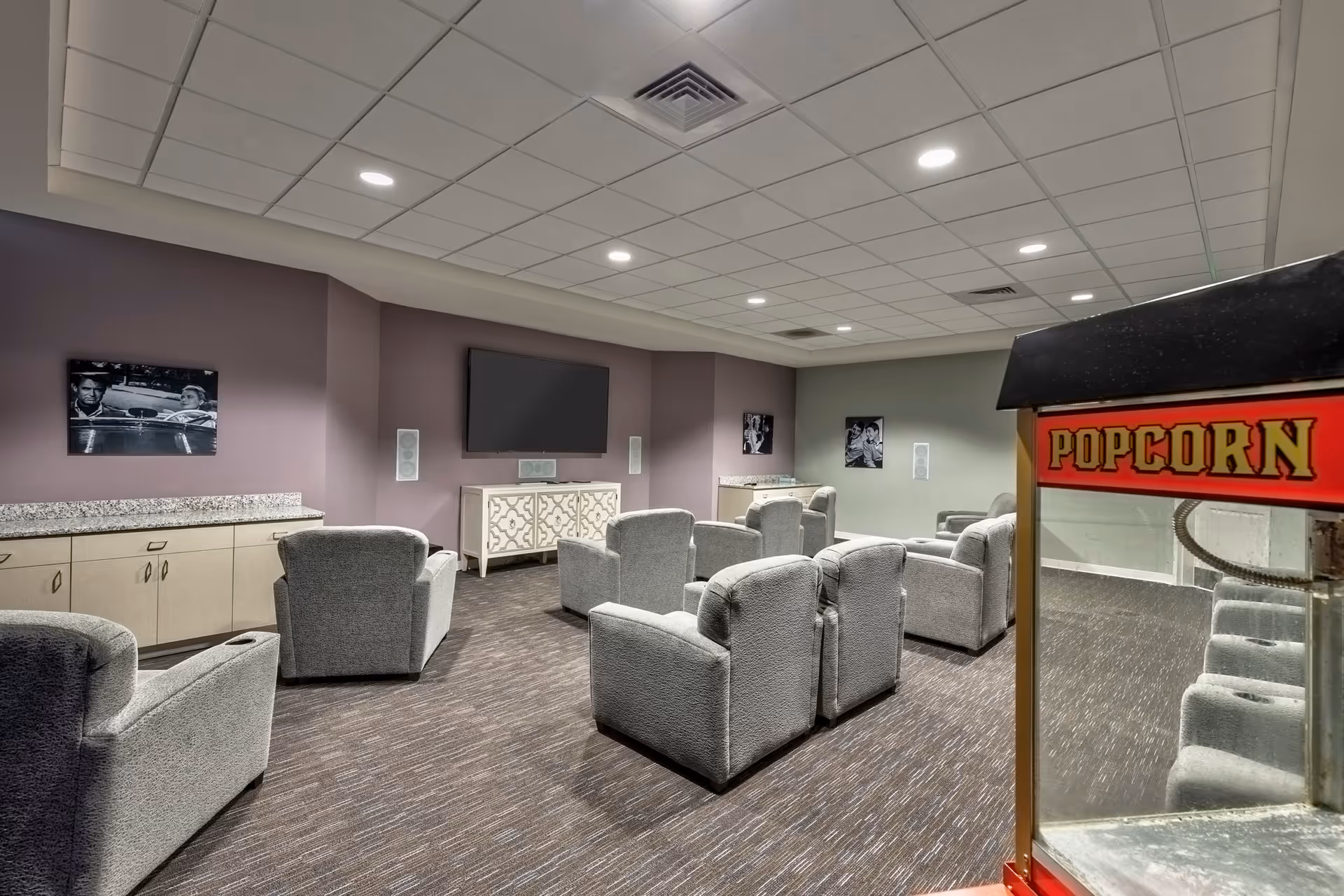 A cozy media room with multiple gray upholstered armchairs arranged in rows facing a large flat-screen TV mounted on a lavender wall. The room has a popcorn machine in the foreground on the right side, carpeted floor, recessed ceiling lights, and black-and-white movie posters on the walls.