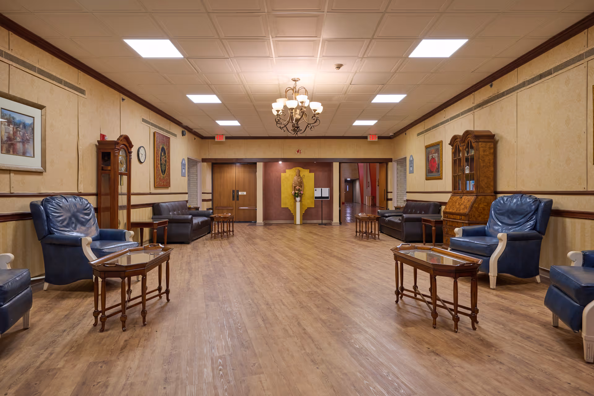 A spacious and well-lit common area with wooden flooring, beige walls, and a white ceiling with recessed lighting and a chandelier. The room is furnished with blue leather armchairs, dark brown leather sofas, wooden side tables with glass tops, a grandfather clock, and a wooden cabinet. Paintings and a clock hang on the walls, and double wooden doors are visible at the far end of the room.