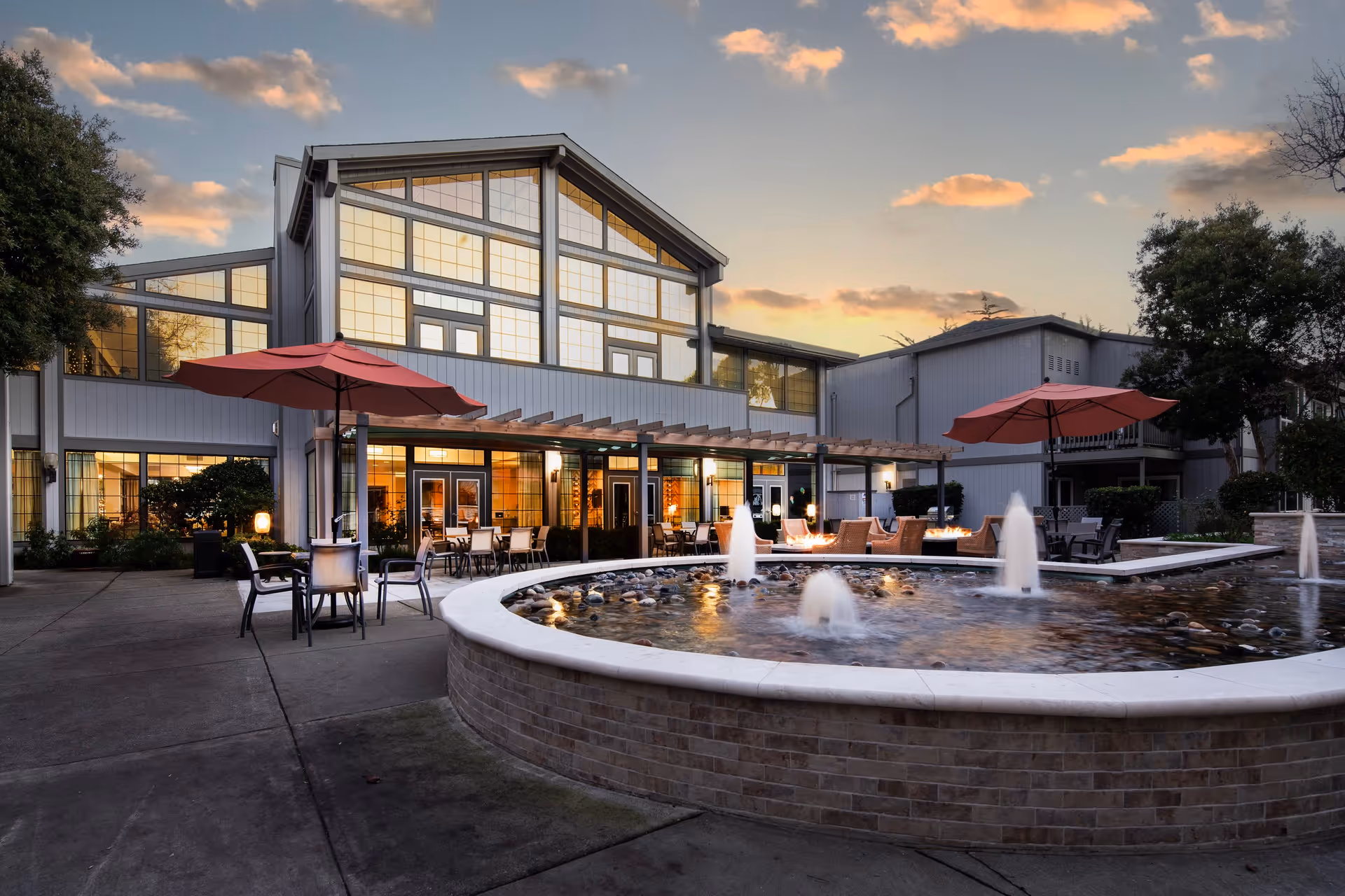 Outdoor patio area at sunset with a large building in the background featuring many windows. The patio has tables and chairs with red umbrellas, and a circular water fountain with multiple small water jets in the foreground.