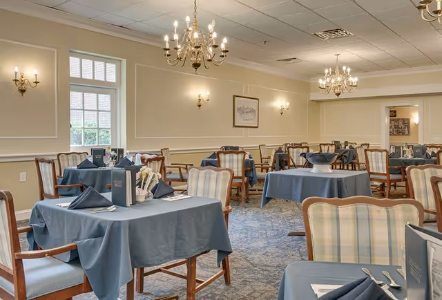 A dining room with multiple tables covered in blue tablecloths, each set with folded napkins, menus, and cutlery. The room features upholstered wooden chairs, chandeliers, wall sconces, framed artwork, and a window letting in natural light.