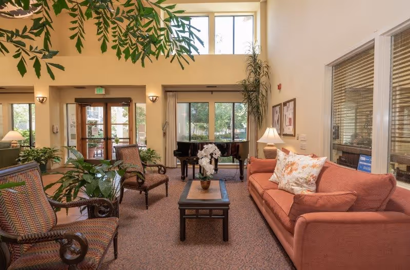 A bright and welcoming senior living facility common area with a coral sofa adorned with floral pillows, two patterned armchairs, a wooden coffee table with a white orchid centerpiece, and a black grand piano near large windows letting in natural light. There are plants and framed artwork on the walls, with a glass door entrance visible in the background.