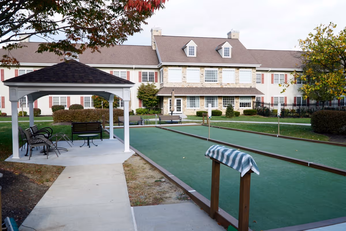 Outdoor bocce ball courts with a white gazebo and seating area in front of a two-story building with stone and beige siding, surrounded by trees and bushes.