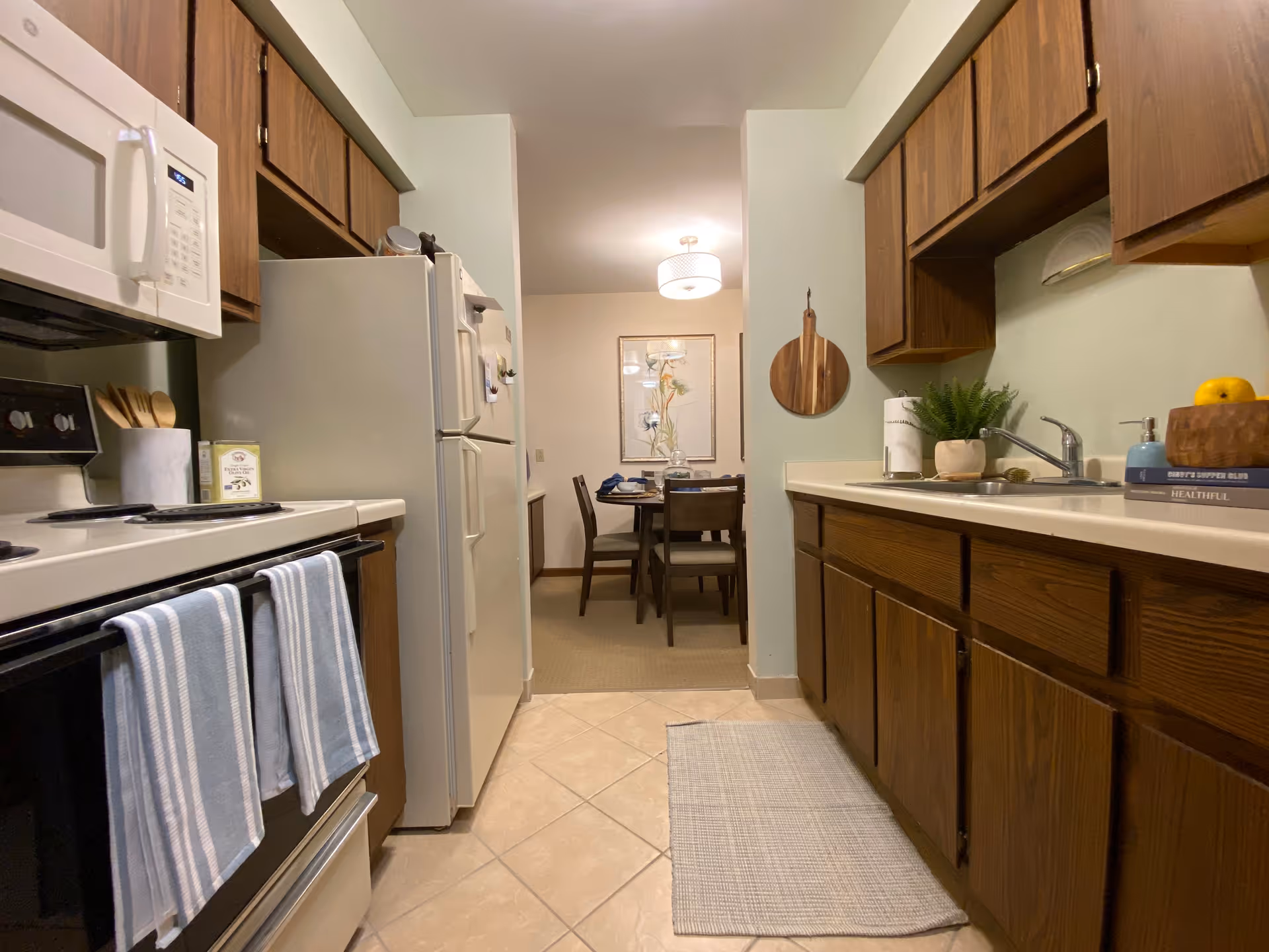 View of a kitchen with wooden cabinets, a white microwave above a stove with two blue-striped towels hanging on the oven door, a white refrigerator, and a sink with a small plant and soap dispenser on the counter. In the background, a dining area with a table and four chairs is visible, along with a framed artwork on the wall and a ceiling light fixture.