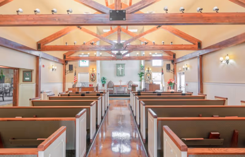 Interior view of a chapel-style gathering room with exposed wooden beams and rows of pews facing an altar.