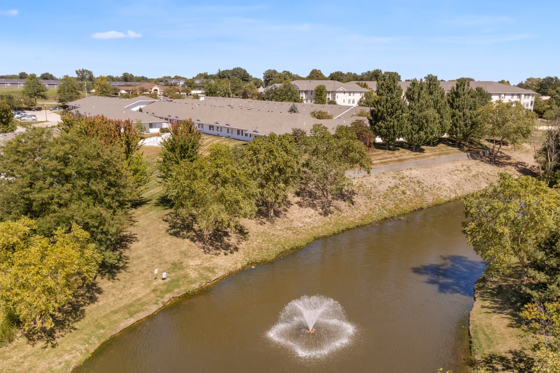 Aerial view of Silvercrest Garner Senior Living facility showing a large building surrounded by trees and greenery, with a pond featuring a water fountain in the foreground under a clear blue sky.