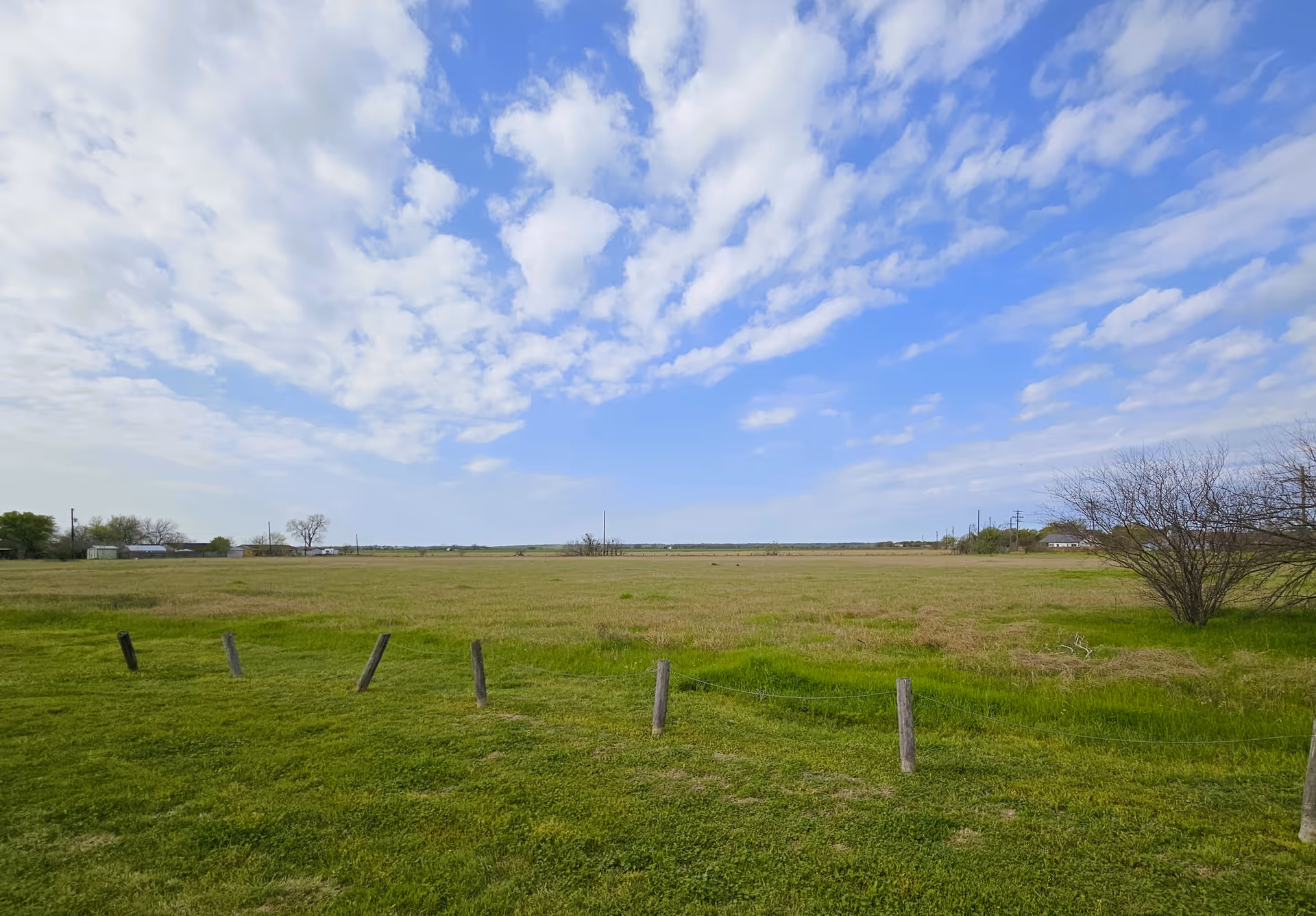A wide open grassy field with a few wooden posts connected by wire in the foreground, a leafless tree on the right side, and a partly cloudy blue sky overhead.