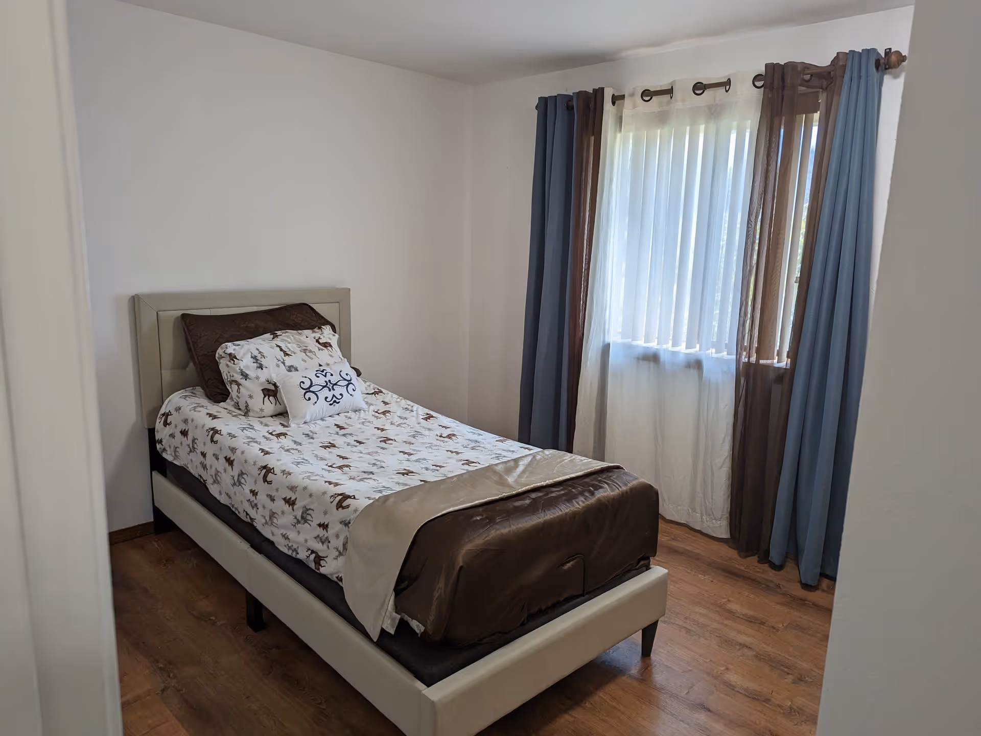 A small bedroom with a single bed featuring a beige upholstered headboard and frame. The bed is made with a white comforter decorated with brown and gray animal and tree patterns, along with several pillows. The room has wooden flooring and a window with sheer white curtains and brown and blue drapes.