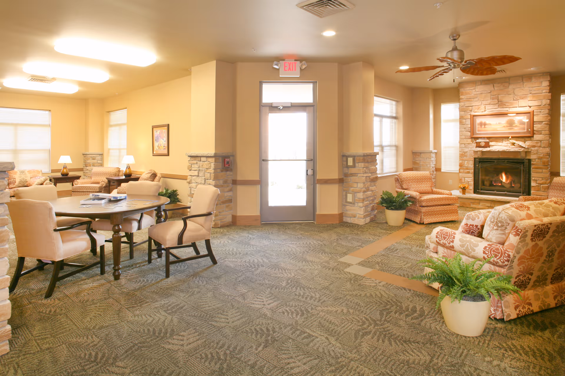 A cozy living room area in a senior living facility with patterned carpet, multiple upholstered chairs and sofas, a round wooden table with chairs, potted plants, a stone fireplace with a framed picture above it, ceiling fan with leaf-shaped blades, and large windows letting in natural light.