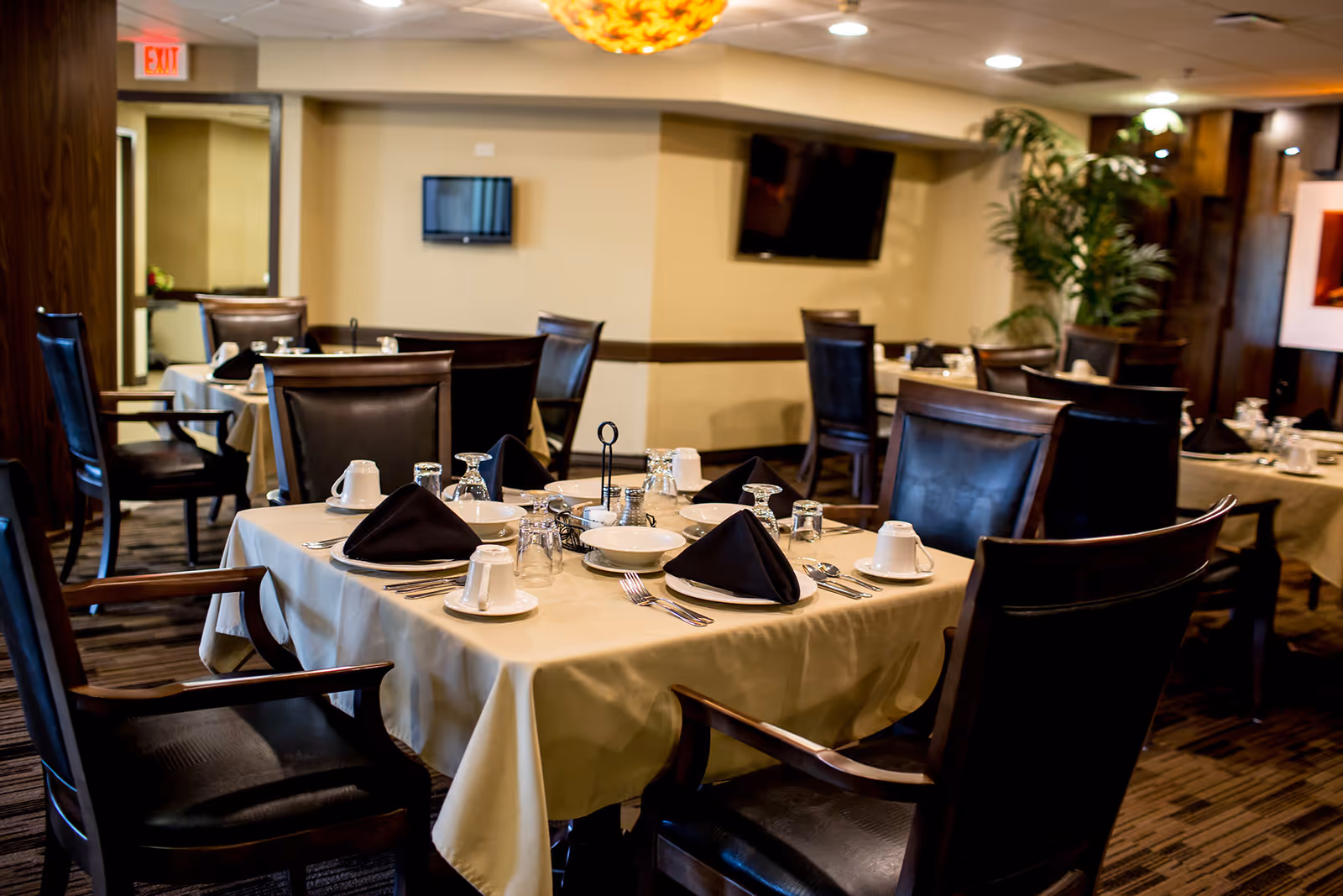 A dining room with tables covered in beige tablecloths, set with black napkins, white cups, plates, and silverware. The room has dark wooden chairs with leather seats and backs, a large decorative light fixture on the ceiling, a flat-screen TV mounted on the wall, and a potted plant in the corner.