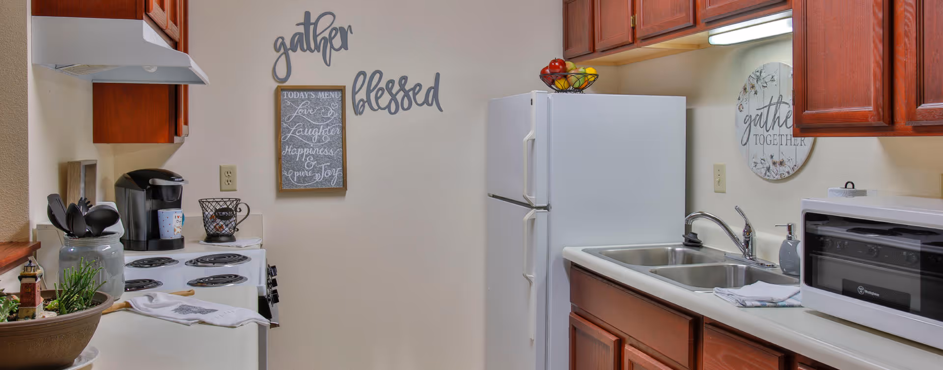 A kitchen area with wooden cabinets, a white refrigerator, a white stove with a coffee maker on top, a double sink, a microwave, and decorative wall signs that say 'gather' and 'blessed'. There is also a small chalkboard with a menu and a bowl of fruit on top of the refrigerator.
