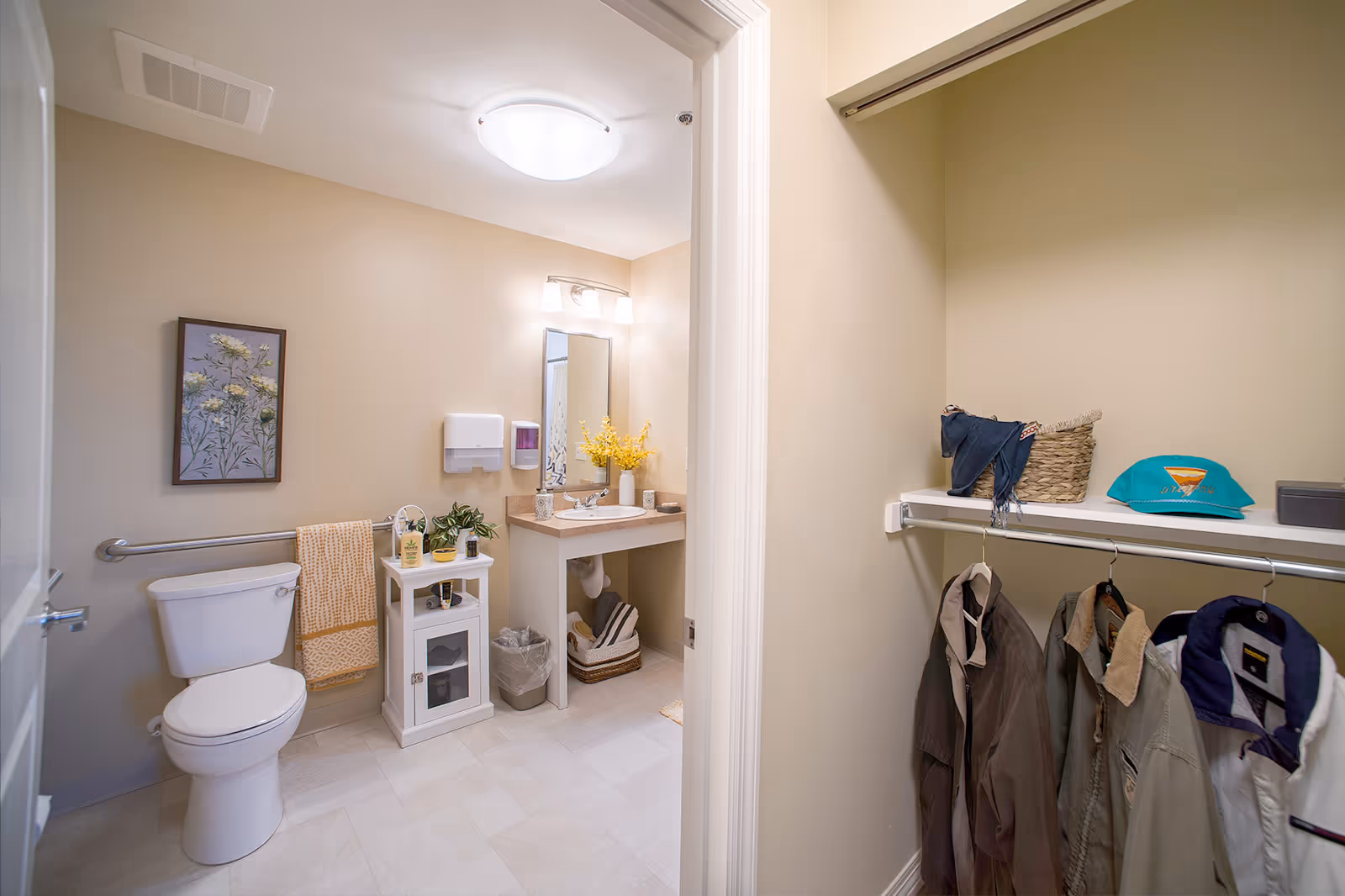 A clean and well-lit bathroom with a white toilet, a small white cabinet with plants and toiletries, a sink with a mirror above it, and a towel hanging on a grab bar. To the right, there is an open closet with several jackets hanging and a shelf holding a basket, a folded scarf, a teal cap, and a small box.