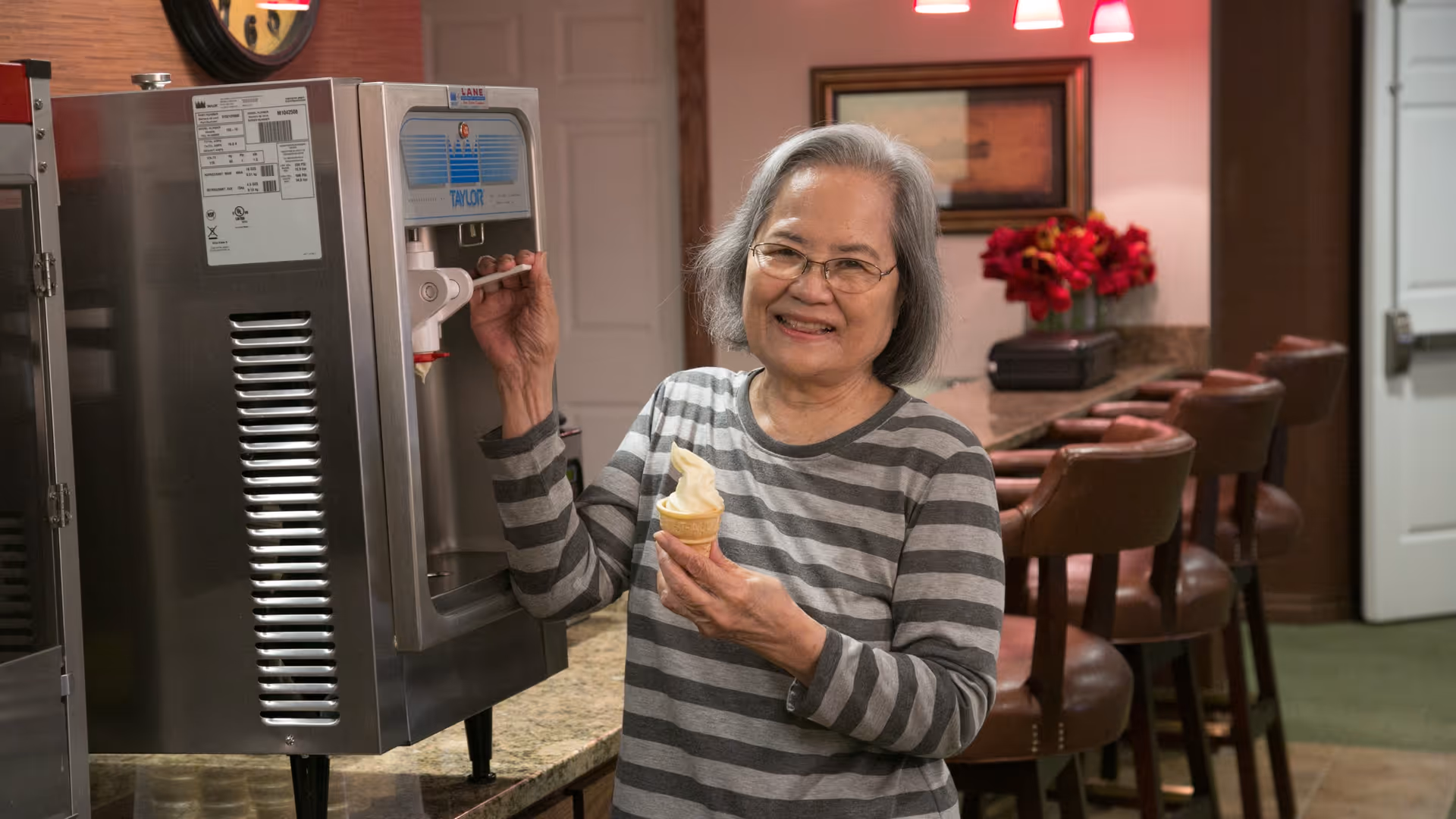 An elderly woman with gray hair and glasses, wearing a gray and white striped shirt, smiling while dispensing soft serve ice cream into a cone from a machine in a cozy room with a counter, bar stools, and red flowers in the background.
