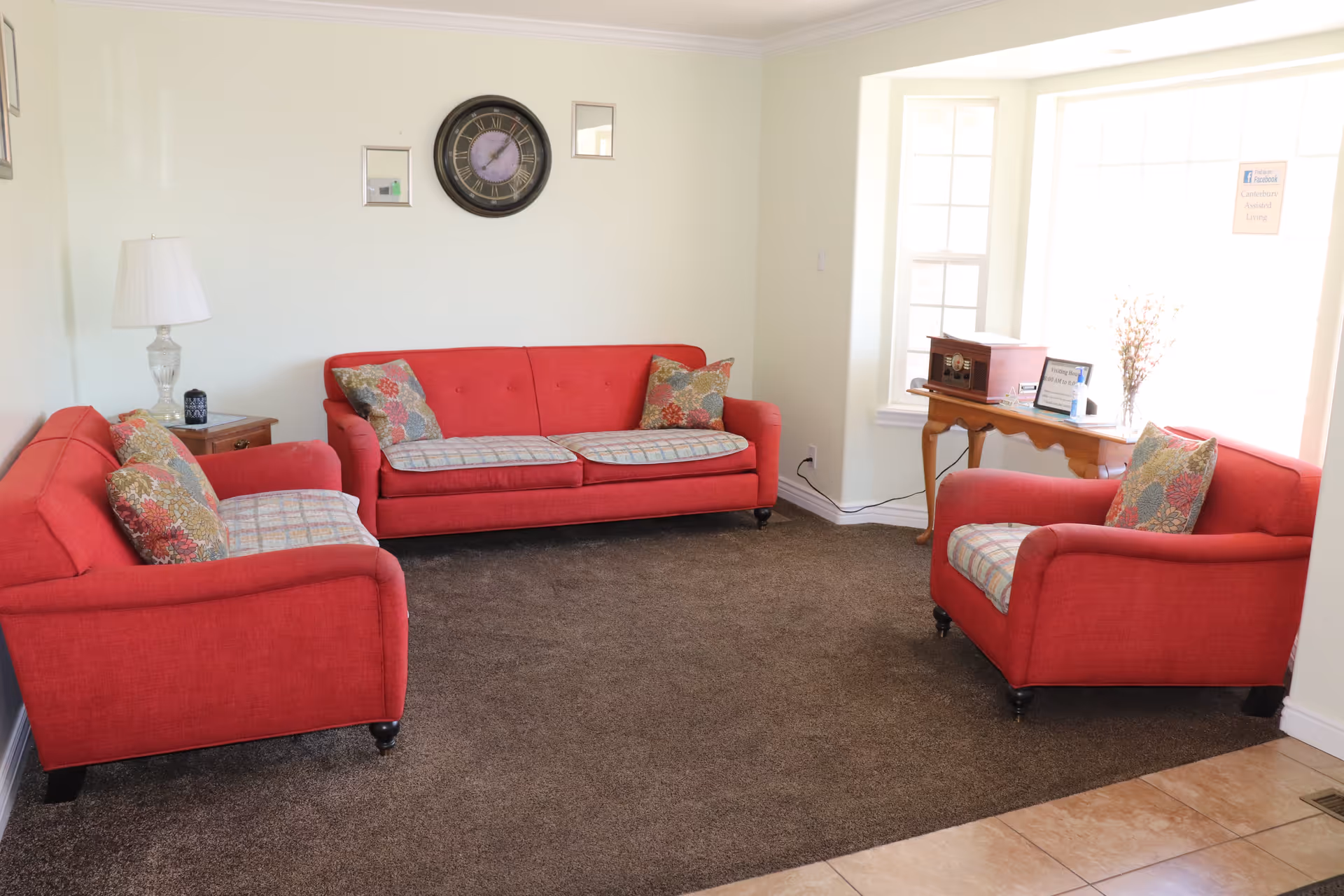 A cozy living room with three red upholstered sofas arranged around a brown carpet. Each sofa has patterned cushions and seat covers. A wooden side table with a glass lamp and a small decorative item is placed beside one sofa. On the wall above the central sofa, there is a large round clock flanked by two small square mirrors. A wooden console table is positioned in front of a large window, holding a vintage-style radio, a framed sign, a hand sanitizer bottle, and a vase with flowers. The room has light-colored walls and tiled flooring near the entrance.