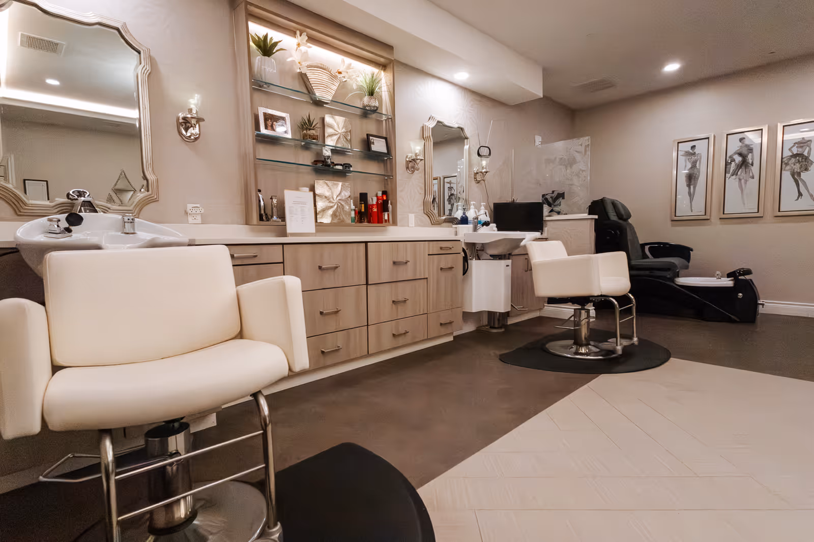 Interior view of a salon area in an assisted living facility featuring two white salon chairs in front of sinks and mirrors, a counter with cabinets and shelves holding decorative items and hair products, and a black pedicure chair in the background. The walls are decorated with framed artwork and the lighting is warm and inviting.