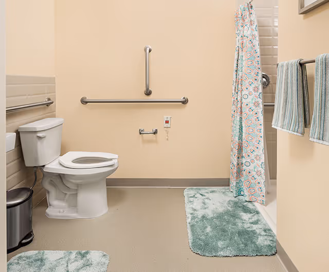 A clean bathroom with a white toilet, grab bars on the walls, a shower with a colorful patterned curtain, green bath mats, and striped towels hanging on a towel rack.