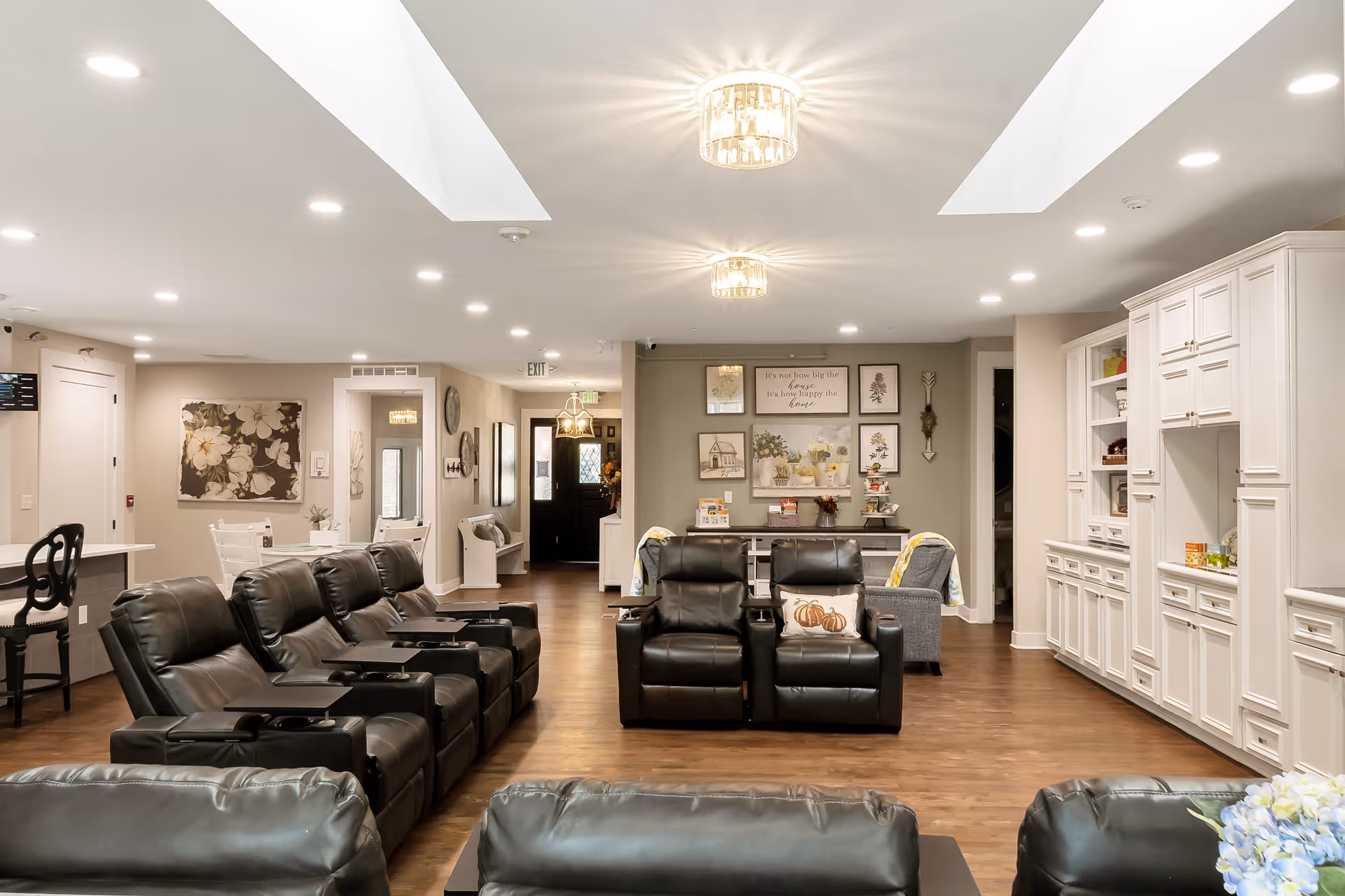 A spacious and well-lit living room area in a senior living facility featuring multiple black leather recliner chairs arranged in rows facing forward. The room has wooden flooring, white built-in cabinets on the right wall, and decorative wall art above a small table at the back. There are two modern ceiling light fixtures and skylights providing natural light. The space also includes a small dining area with white chairs and a counter with black chairs on the left side.