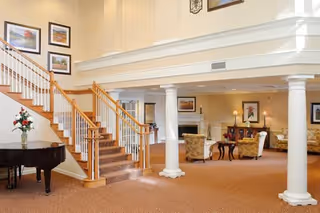 Spacious senior living facility common area with a grand staircase featuring wooden handrails and carpeted steps. The room has beige walls and carpet, white columns, a black grand piano with a flower arrangement on top, framed artwork on the walls, and comfortable seating including armchairs and a sofa near a fireplace.