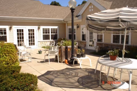 Outdoor patio area at a senior living facility with white metal chairs and tables, a striped umbrella providing shade, potted plants on tables, a lamp post in the center, and beige building walls with multiple windows and doors in the background.