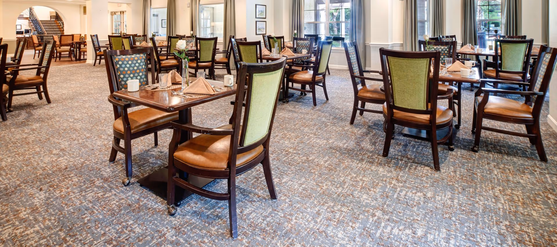 Dining room with multiple wooden tables and upholstered chairs arranged on patterned carpet, each table set with napkins and cups.