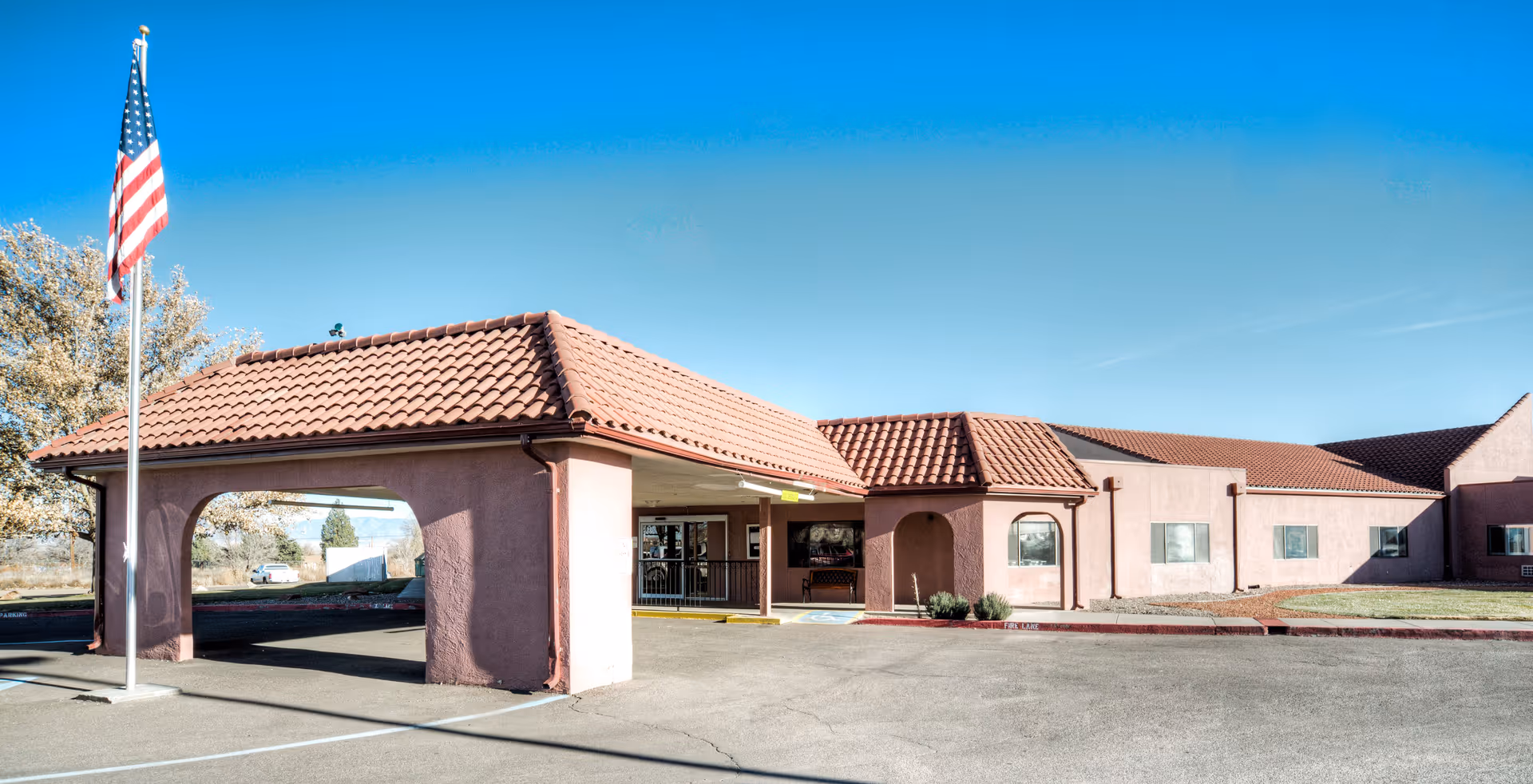 Exterior view of Belen Meadows Healthcare and Rehabilitation Center showing a single-story building with a red tiled roof and a covered entrance. An American flag is flying on a flagpole near the entrance. The sky is clear and blue.