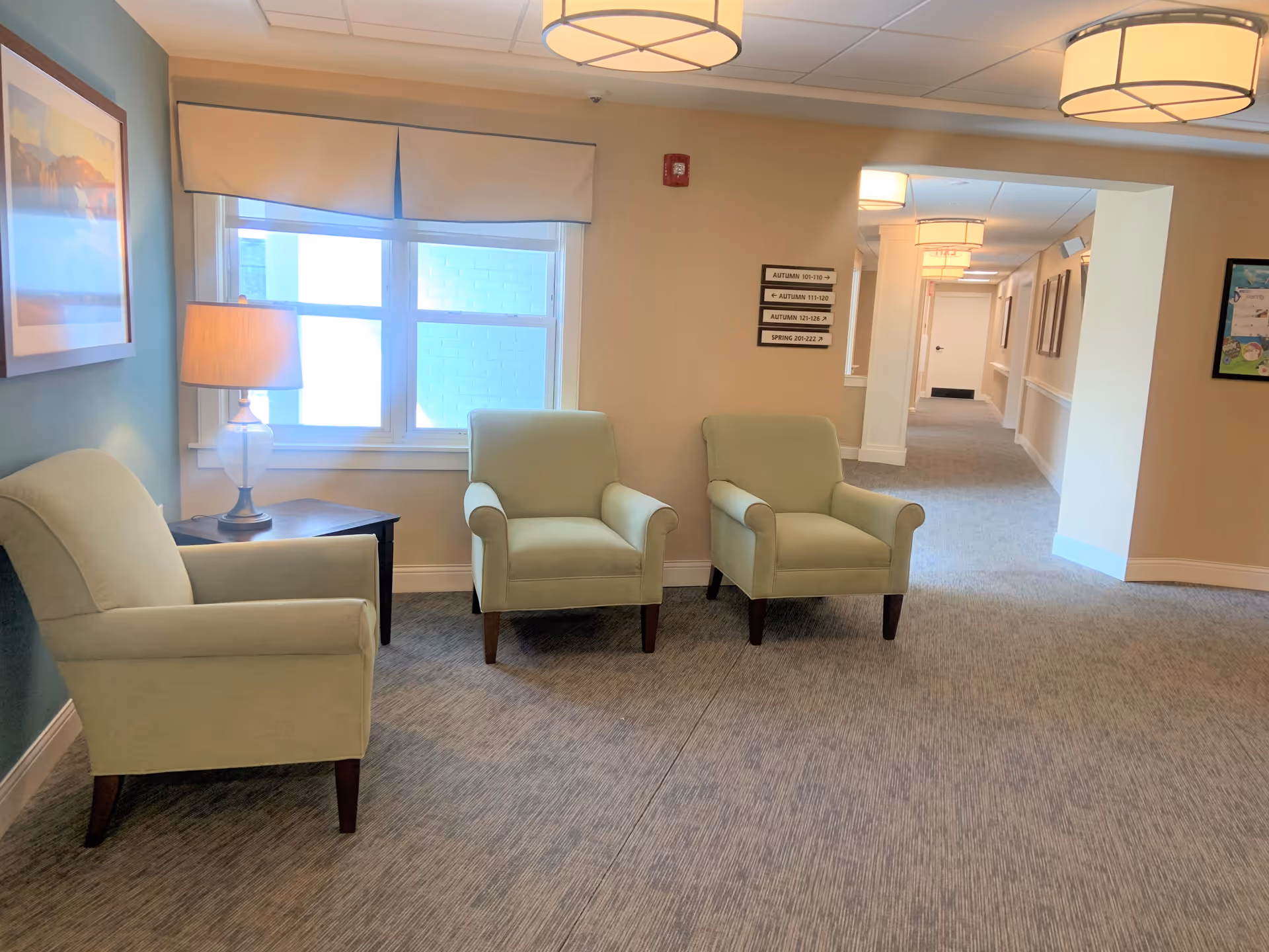 A cozy seating area in a senior living facility with three light green armchairs arranged near a window with a beige valance. A table with a lamp is next to one chair, and a hallway with directional signs is visible in the background. The walls are painted in soft beige and teal colors, and the carpet is gray.