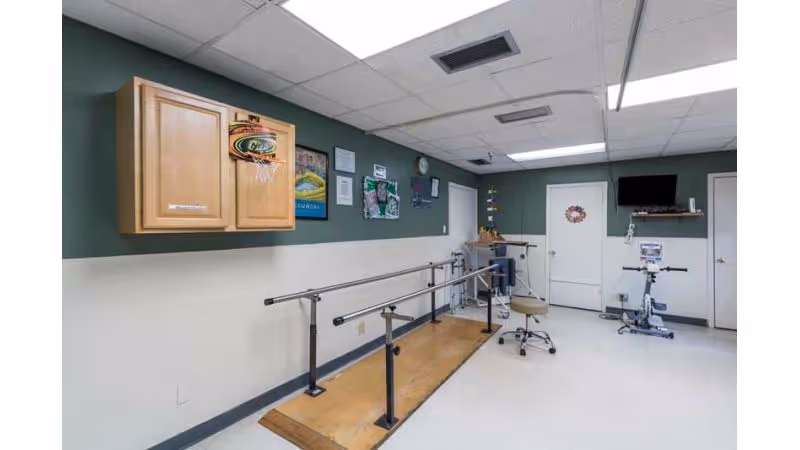 A rehabilitation room with parallel bars for walking exercises, a small basketball hoop mounted on a wooden cabinet, a stationary exercise bike, a rolling stool, and various posters and equipment on the walls.