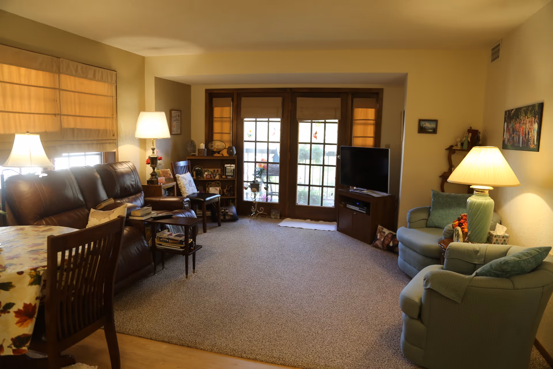A cozy living room with a brown leather sofa, two green armchairs, a wooden dining table with chairs, a TV on a wooden stand, and a bookshelf. The room has beige walls, carpeted floor, and large glass doors with wooden frames leading outside. Several lamps provide warm lighting, and there are decorative items and pictures on the walls and shelves.
