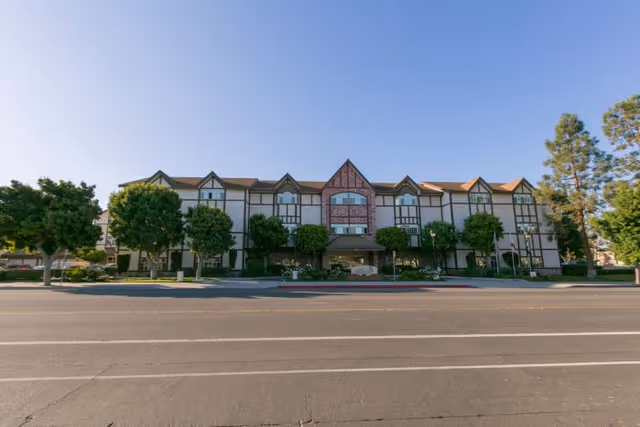 Front exterior of a three-story Tudor-style assisted living building with trees and a road in front.